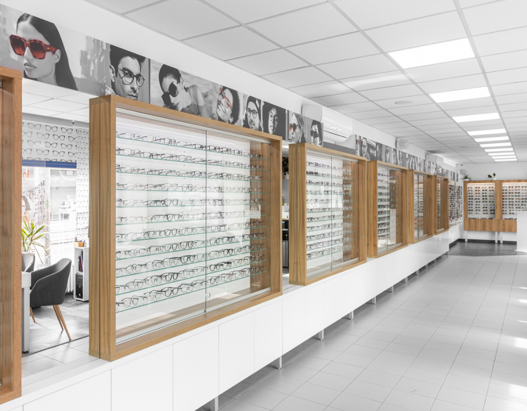 Interior of an optics store with wooden-framed glass display cases filled with eyeglasses along a white-tiled aisle image-wide.